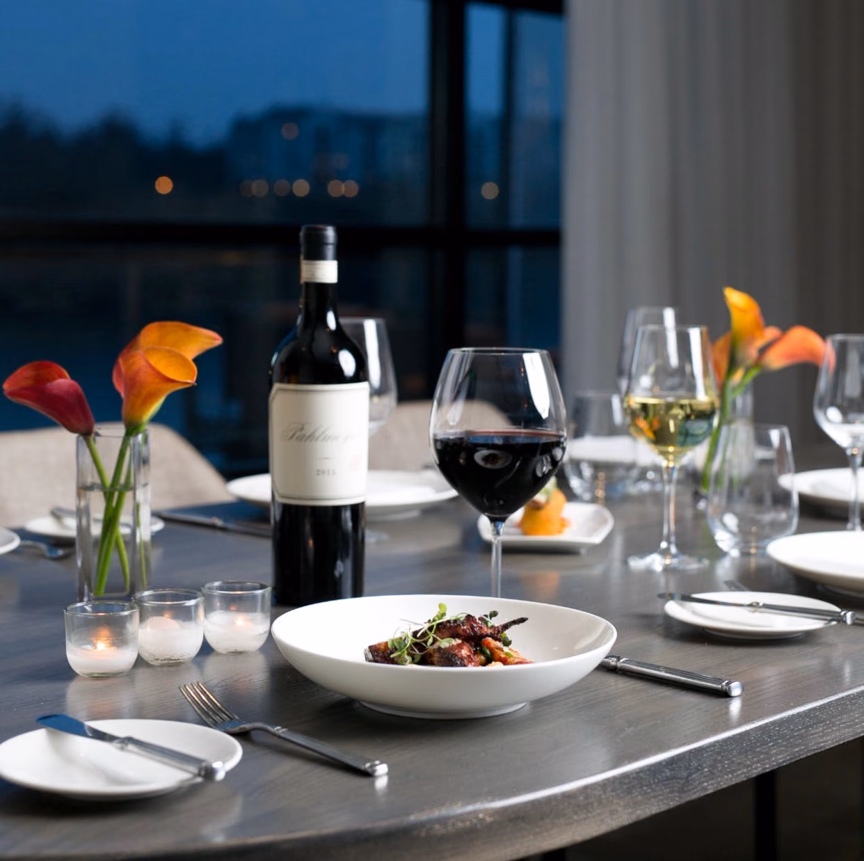 A candlelit dinner at Citron Baltimore, a plated dish with red and white wine and the Quarry Lake view glowing through the windows at dusk